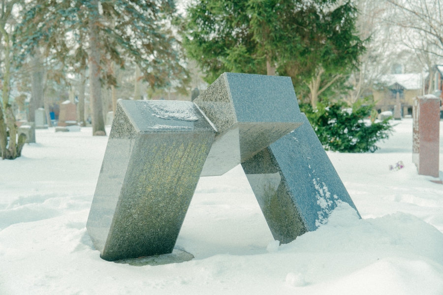 Abstract stone sculpture composed of angled geometric slabs rising from the ground in a snow‑covered cemetery. Trees and surrounding grave markers are visible in the winter landscape.