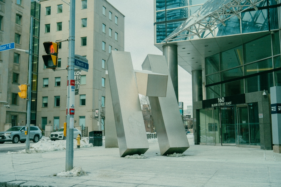 Large abstract metal sculpture made of tall, angular geometric forms installed on a city sidewalk. Modern office buildings, glass façades, and street signage surround the sculpture in an urban downtown setting.