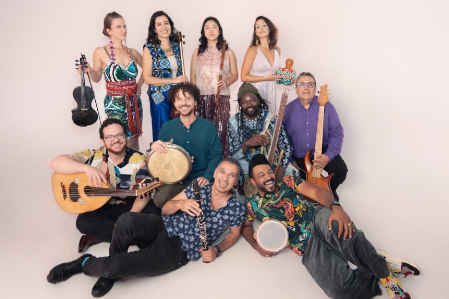 Group of musicians posing together with various instruments, including drums, violin, and guitar, against a plain background.