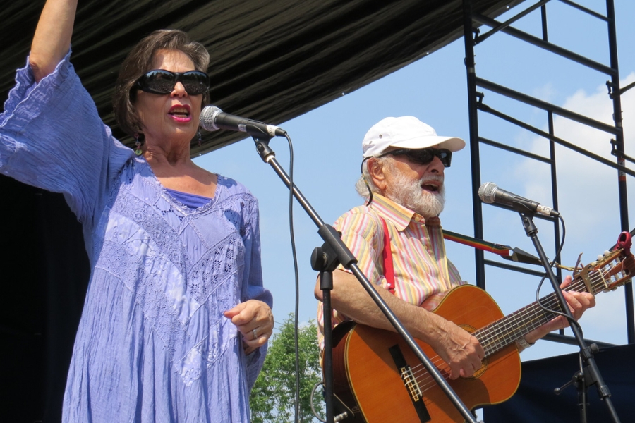 Two performers stand on an outdoor stage; one is singing into a microphone with an arm raised, while the other plays an acoustic guitar beside an additional microphone stand. A canopy and metal stage scaffolding are visible overhead against a sunny sky.