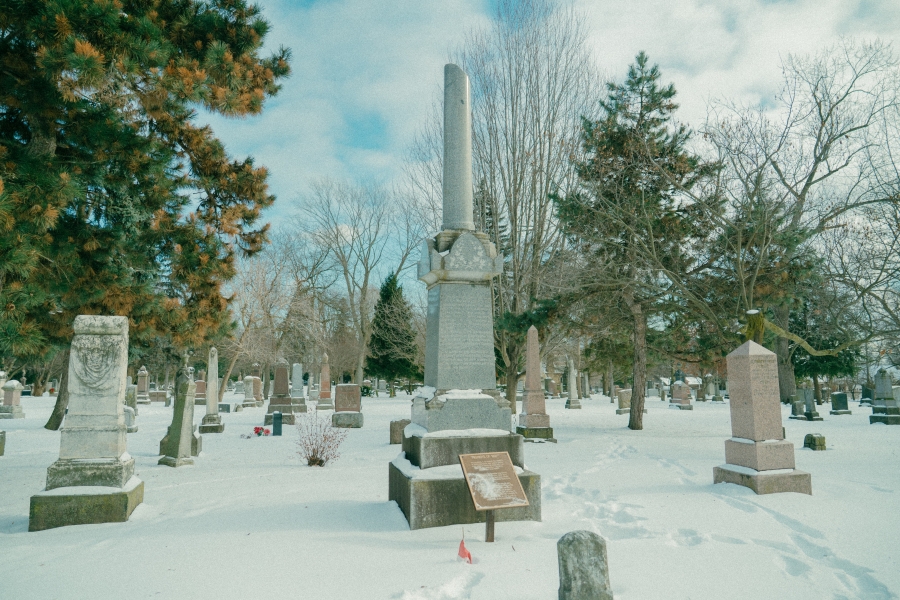 Snow‑covered cemetery landscape featuring the Lount and Matthews monument, a tall stone memorial with a column rising from a stepped base. The monument stands among surrounding headstones and leafless trees in a winter setting, with an interpretive plaque placed in front.