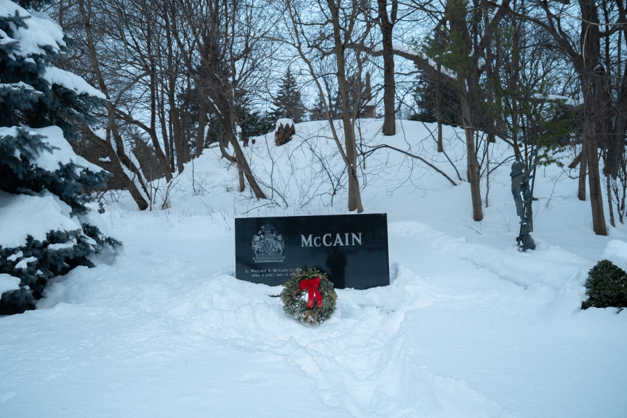 Snow‑covered cemetery scene featuring the McCain family marker, a low black granite headstone set against a wooded hillside. A green memorial wreath with a red bow rests in front of the stone, with trees and snow‑covered ground surrounding the grave in winter.