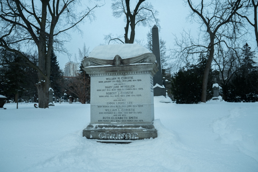 Snow‑covered cemetery scene featuring the McCain family marker, a low black granite headstone set against a wooded hillside. A green memorial wreath with a red bow rests in front of the stone, with trees and snow‑covered ground surrounding the grave in winter.