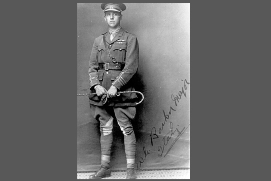 A full‑length black‑and‑white portrait of William George Barker standing in front of a plain studio backdrop. The officer is dressed in early 20th‑century military attire with a belted jacket, medals on the chest, tall boots, and a cap. He holds a riding crop or swagger stick in both hands, positioned in front of him. A handwritten note or signature is visible along the lower right side of the photograph.