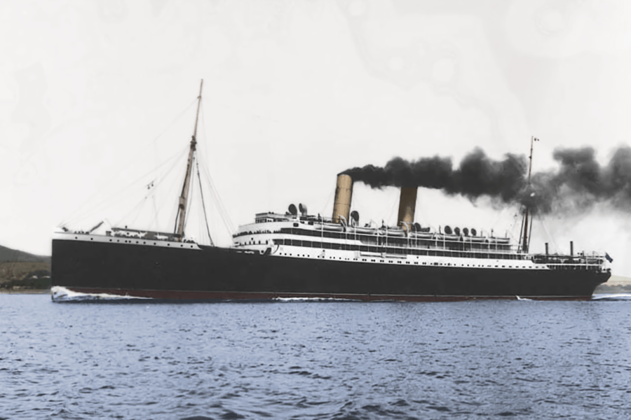 A large early‑20th‑century ocean liner with a black hull and white superstructure steams across calm water, emitting dark smoke from multiple funnels, viewed broadside against a pale sky and distant shoreline.