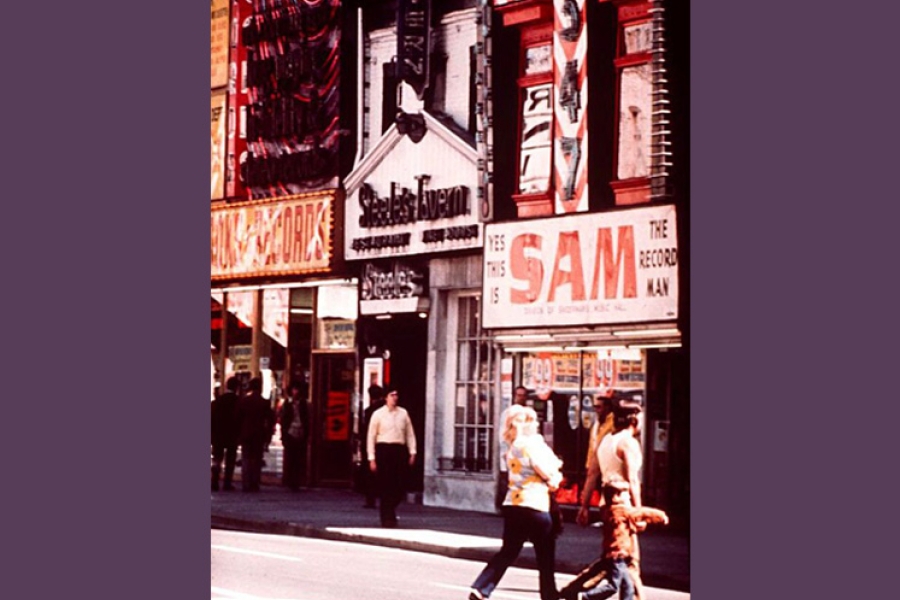 A street scene featuring colorful storefronts, including Sam the Record Man and Steele’s Tavern. Pedestrians walk across the street in the foreground, while others stand near the shop entrances. Bright signage and busy façades create a lively urban atmosphere.