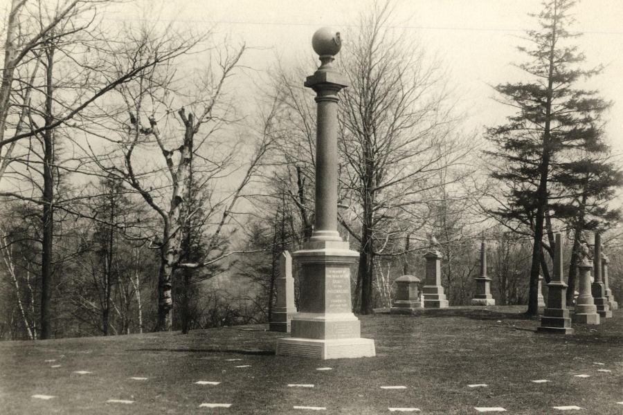 A black-and-white photograph of the Freemasons’ burial plot in a historic cemetery, featuring a tall stone column monument topped with a sphere in the foreground. Rows of smaller gravestones and monuments are arranged across a gently sloping grassy landscape, with leafless trees in the background, suggesting a formal memorial setting in late fall or early spring.