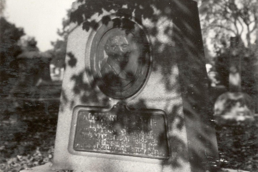 A black‑and‑white photograph of a stone grave monument standing outdoors among fallen leaves. The monument is tall with a sloped top and features an oval sculpted portrait relief of a man on its front. Beneath the portrait is an inscribed plaque, though the text is difficult to read due to shadows from nearby tree branches. Trees and other headstones are visible in the background.