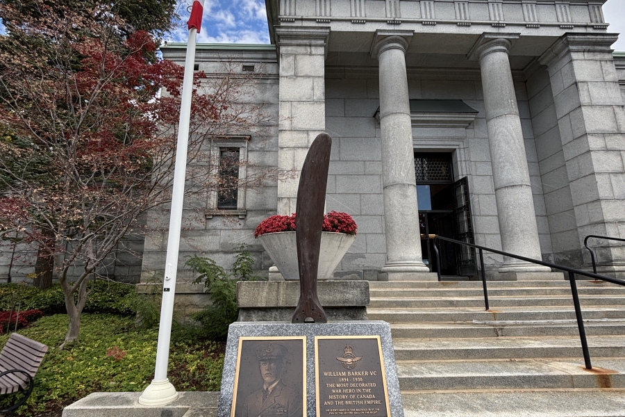 Outdoor photograph of a stone memorial set at the base of wide steps leading to a classical stone building with tall columns and a triangular pediment. In the foreground, a granite monument holds two bronze plaques and a vertical sculptural element shaped like an aircraft propeller. A Canadian flag stands on a pole to the left, with trees and landscaped greenery surrounding the area. The sky above is partly cloudy, and the scene conveys a formal, commemorative setting connected to history and remembrance.