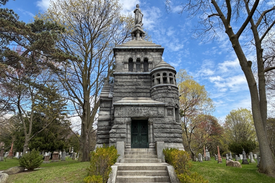 Stone mausoleum with steps, arched windows, and a statue on top, surrounded by trees and gravestones.