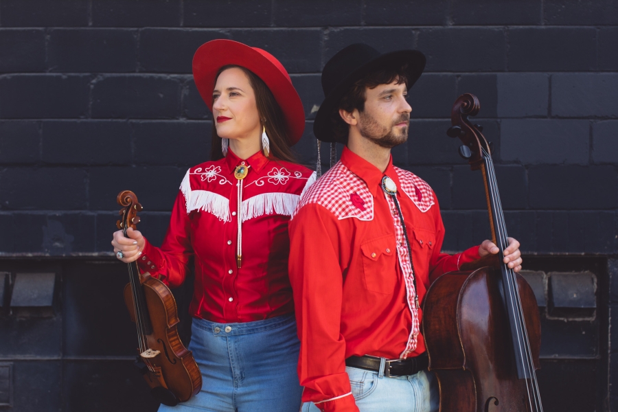 Two musicians in red western-style outfits standing back-to-back, holding a violin and a cello.