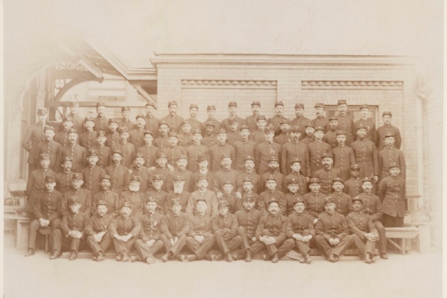 A large group of Toronto uniformed postal workers, including Albert Jackson, posed in several rows outside a brick building, with men seated and standing in formal arrangement, wearing matching caps and coats in a late‑19th‑century group portrait.