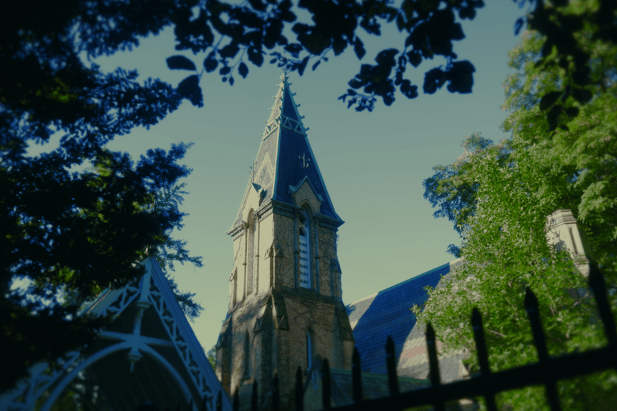 View of the Toronto Crematorium Chapel, a Gothic‑style stone building with a tall central tower, seen through tree branches and a wrought‑iron fence in a leafy, sunlit setting.