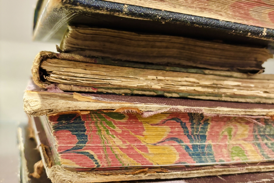 Stack of worn, old hardcover books with frayed edges and faded patterned covers, showing signs of heavy use and age, resting on a flat surface.