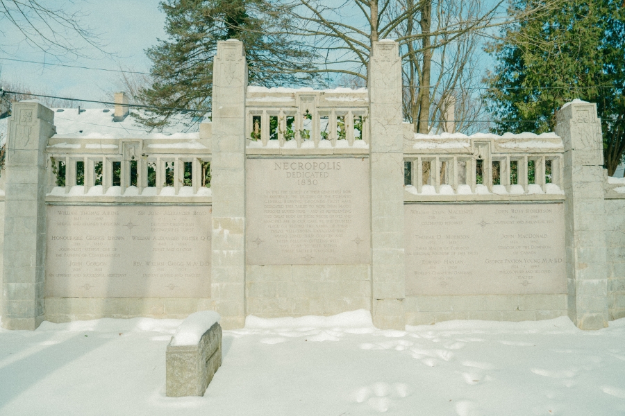 Snow‑covered stone dedication wall at the Toronto Necropolis, featuring engraved memorial panels framed by vertical pillars and decorative stonework. Trees rise behind the wall, and a low stone marker sits in the foreground within the winter landscape.