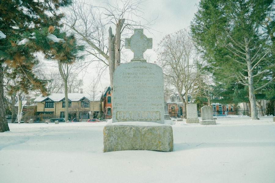 Snow‑covered cemetery scene featuring the grave marker of Ned Hanlan, a tall stone headstone topped with a cross and set on a rectangular base. The monument stands among trees and surrounding grave markers, with residential buildings visible in the background during winter.