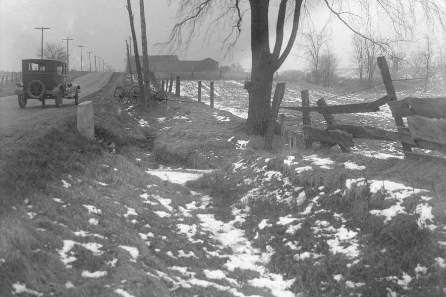 A black-and-white photograph of an undeveloped rural roadside in winter, with patches of snow scattered across uneven ground in the foreground. A dirt road runs along the left side of the image, with an early automobile parked near a line of utility poles. Wooden fence posts, a leafless tree, and open fields extend into the distance, with farm buildings faintly visible on the horizon.