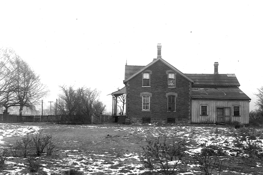 A black-and-white photograph of a rural brick farmhouse with a gabled roof and a small wooden addition on one side, set on an open field with patches of snow on the ground. Leafless trees, a fence line, and utility poles surround the property, suggesting a winter scene in an early 20th-century rural setting.