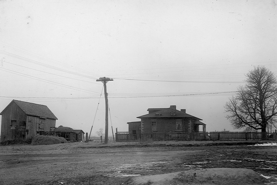 A black-and-white photograph of a small rural settlement showing a one-storey house with a hip roof and front porch enclosed by a wooden fence, alongside a large wooden barn and outbuildings. A dirt road runs across the foreground, with utility poles and overhead wires cutting across the scene, leafless trees nearby, and patches of snow on the ground, suggesting a winter landscape from the early 20th century.