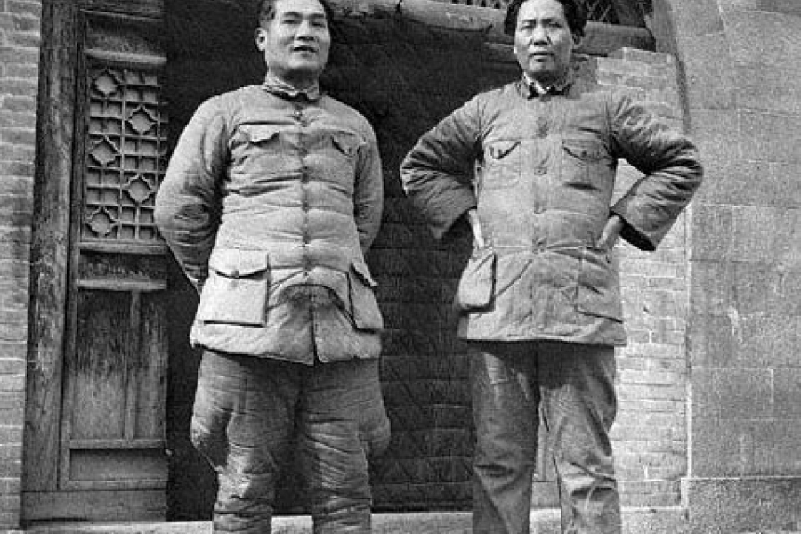 Black‑and‑white photograph of two people wearing padded military jackets, standing side by side in front of a stone building with a carved emblem above the doorway. Both figures stand confidently with hands positioned at their sides or on their hips.