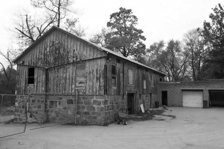 Black and white photograph of the barn structure in the Pine Hills Cemetery maintenance yard. There are tall trees behind, some are bare and others are coniferous.