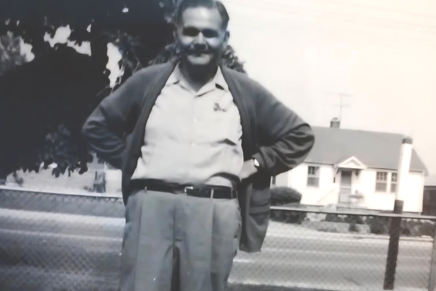 Black and white photo of Mel Chandler standing with his hands on his hips. He is wearing a light-coloured button-down dress shirt, belted dress pants, and a cardigan. Behind him is a large leafy tree and a house across the road. Behind him are the gates to Pine Hills Cemetery, at the Kennedy side exit.