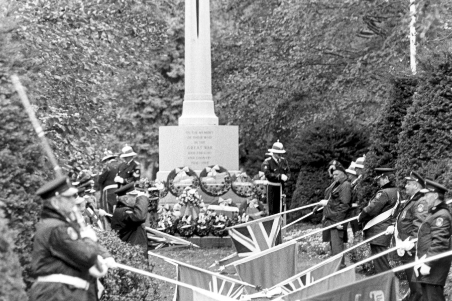 Black-and-white photo of a ceremonial gathering at a memorial cross, with uniformed participants standing in formation and flags displayed in front of the monument, surrounded by trees.
