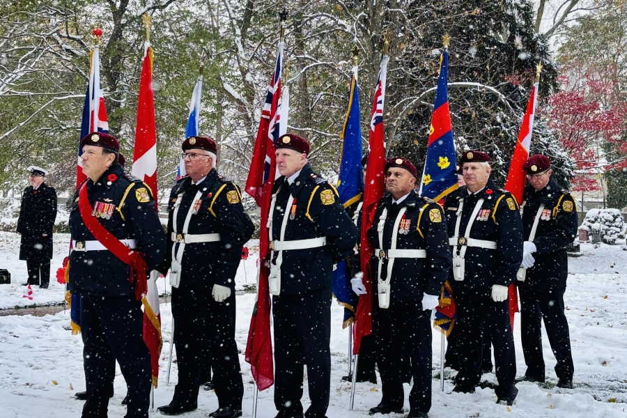 Six servicemen carrying flags, standing with their arms to the side. There is snow on the ground and the trees in the background have snow on their leaves and needles.