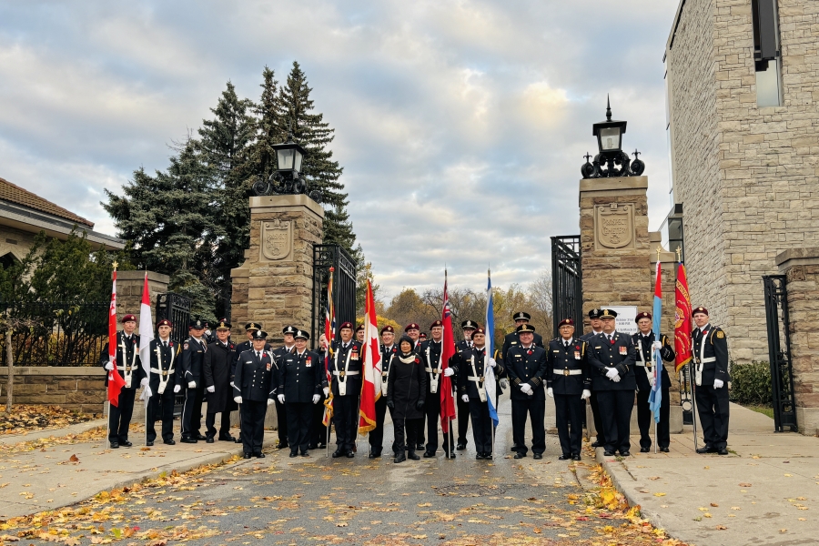 About 25 people gather between two stone pillars at the gate of Prospect Cemetery. There are a few flags held by members of HCMS York. The mayor of Toronto, Olivia Chow is in the centre of the photo. There are yellow leaves on the road leading into the cemetery, and the sky is cloudy with some blue sky peeking through.
