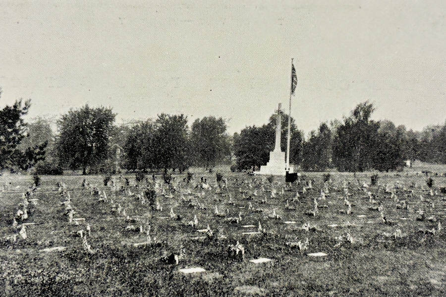Historic black-and-white photograph of a veterans’ burial plot with rows of small grave markers spread across a grassy field, centered on a tall Cross of Sacrifice with a flag. Mature trees line the background