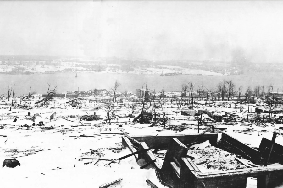 Black‑and‑white photograph of widespread destruction along a waterfront following an explosion. Snow‑covered ground is strewn with debris and collapsed structures, with leafless trees and damaged buildings visible across the landscape and the harbour in the distance.