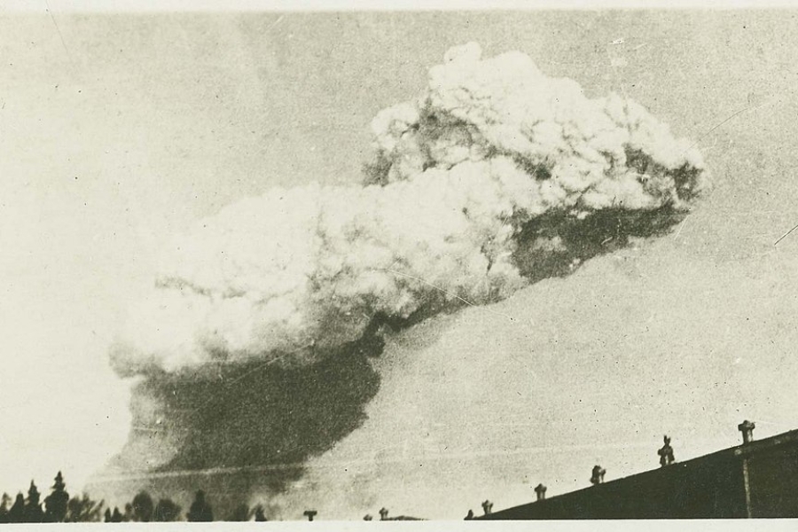 Historic black‑and‑white photograph showing a massive explosion cloud rising into the sky above buildings and rooftops. The cloud billows outward in dense layers, dominating the horizon while silhouetted structures and figures appear along the lower edge of the image.
