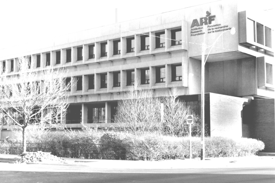 A black‑and‑white exterior view of the Addictions Research Foundation where Eleanor worked as a therapist. It is a mid‑century institutional building with a concrete façade and rows of rectangular windows, set back from a city street with bare trees and low shrubs in the foreground. The photo was taken in the 1990s.