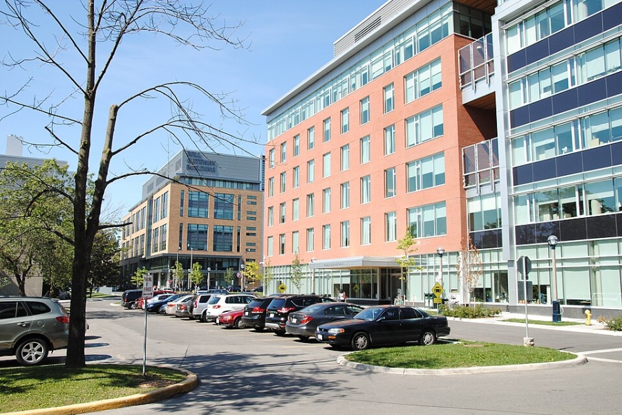 Exterior view of a modern multi‑storey institutional building with red brick and glass façades, large windows, and a stepped design, fronted by a parking lot with cars, young trees, and pedestrian walkways under a clear blue sky.