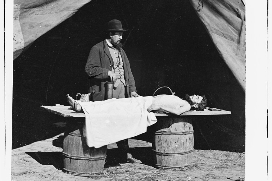 Black‑and‑white historical photograph showing an embalming surgeon standing beside a covered body laid on a wooden table supported by barrels, set outdoors beneath a canvas tent. Medical equipment and containers rest on the table, and the ground beneath is bare earth, reflecting a field mortuary setting.