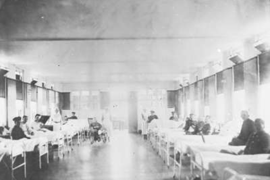 Black‑and‑white photograph of a long hospital ward with rows of metal‑framed beds on either side, occupied by recovering soldiers, with nurses attending patients beneath large windows lining the room.