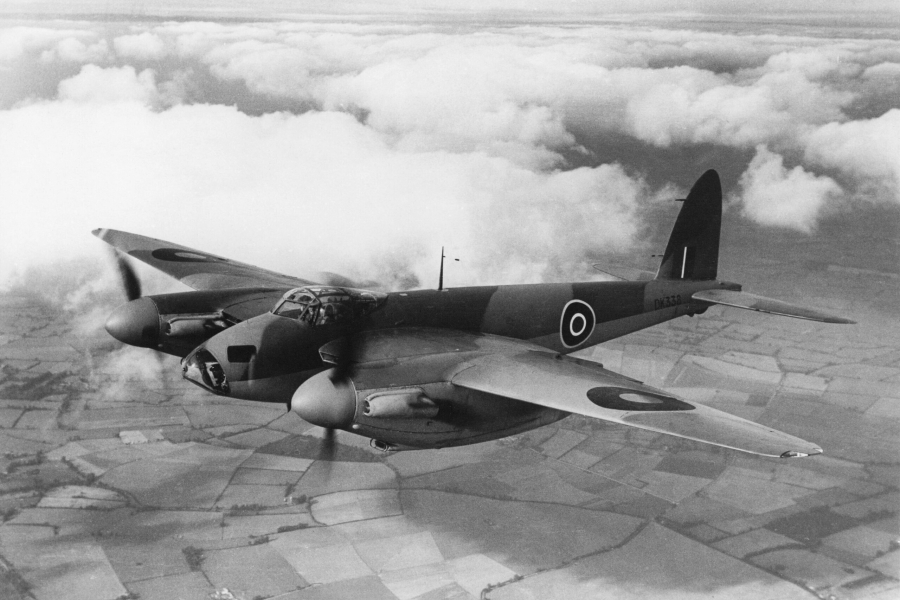 Black‑and‑white photograph of a military aircraft flying above patchwork farmland and clouds. The twin‑engine plane bears Royal Air Force roundels on its wings and fuselage and is shown in flight from a side angle, with clouds stretching across the horizon below.