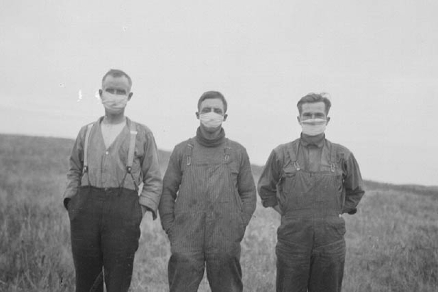 Black-and-white photograph of three young people standing side by side in a grassy field. They are wearing heavy work clothes with long sleeves and overalls, suggesting farm or manual labor. All three of them are wearing medical masks. The field slopes gently behind them, with open land and sky in the background. The scene has a stark, historical feel, with simple clothing and a rural setting.