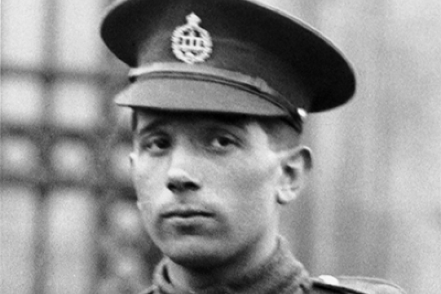 A historical black‑and‑white photograph of a uniformed military figure wearing a peaked cap with an insignia. The person is dressed in a formal service uniform, standing outdoors in front of a softly blurred background with architectural elements.