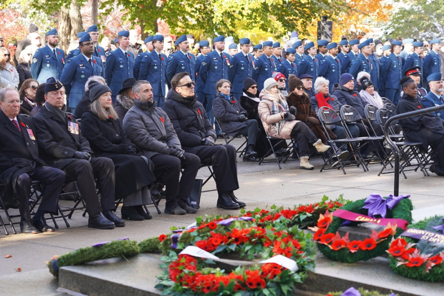 People seated at an outdoor memorial ceremony with wreaths displayed in the foreground.
