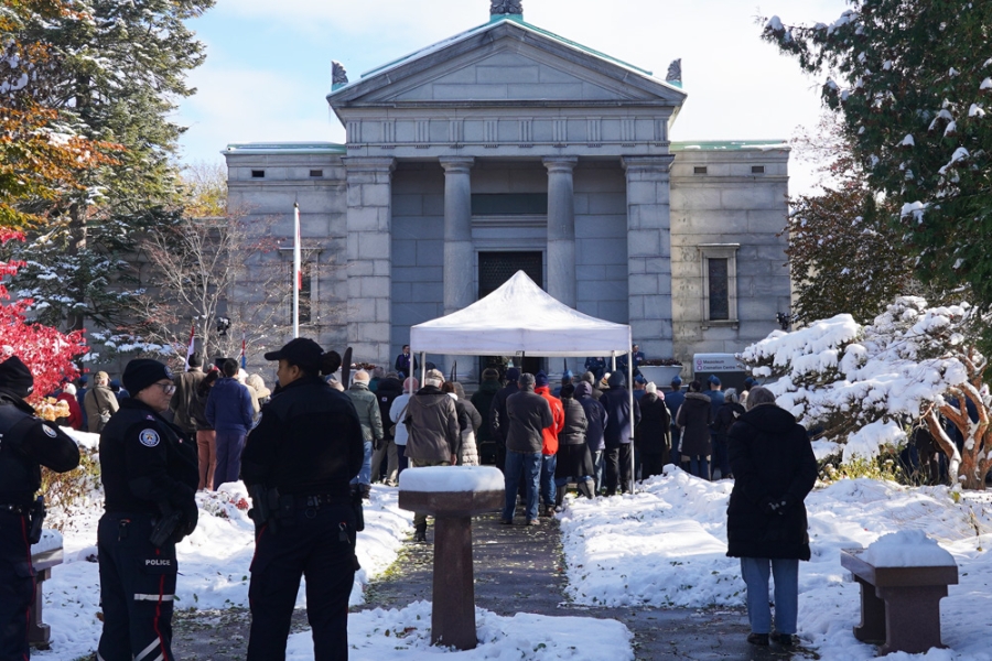 A crowd gathers outside a stone building during a winter memorial event, with snow lining the walkway.
