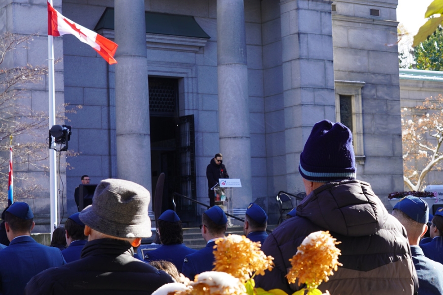A speaker stands at a podium outside a stone building during a memorial ceremony, with attendees gathered in the foreground and a Canadian flag flying nearby.