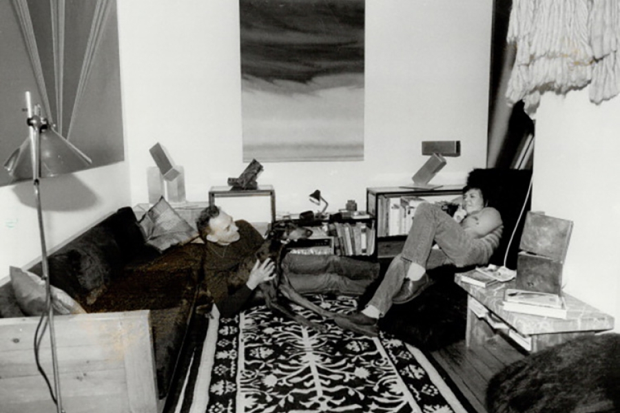 Two people sitting on the floor in a living room, talking beside a sofa and patterned rug with books and artwork nearby.