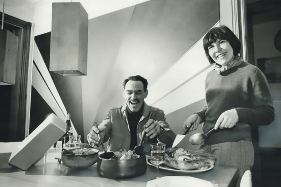 Woman serves food as a man laughs at a dining table in a modern kitchen.