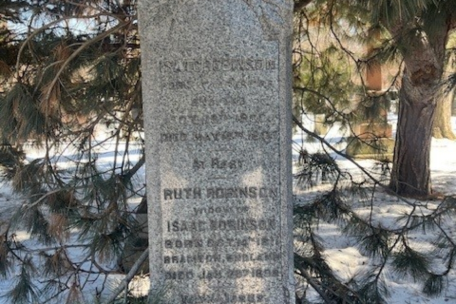 Weathered gray stone grave marker with an arched top, partially obscured by pine tree branches, standing in a snowy cemetery; the engraved inscription is faint and difficult to read.