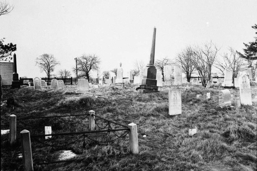 Black and white photo of a burial ground. There are some trees in the background, and it appears to be late autumn. 