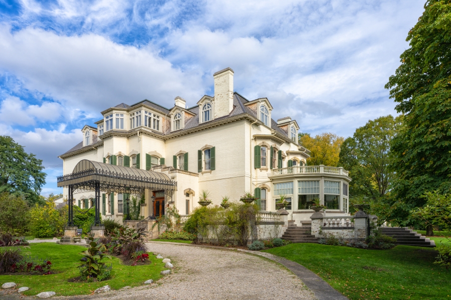 Exterior view of Spadina House, a historic two‑and‑a‑half‑storey mansion with cream‑coloured walls, green shutters, and a mansard roof, featuring a covered entrance, bay windows, and landscaped gardens with a curved driveway and mature trees under a partly cloudy sky.