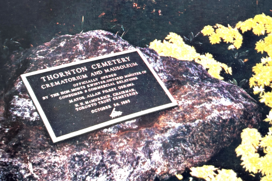 A commemorative plaque mounted on a large rock at Thornton Cemetery marks the opening of the crematorium and mausoleum, surrounded by yellow flowers and fallen leaves outdoors.