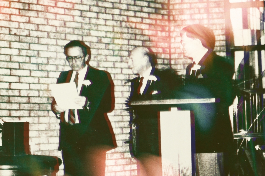 Three men in formal suits stand at a podium in front of a brick wall. A letter from Ontario Premier David Peterson is being read by the Honourable Monte Kwinter, just prior to being sealed in a time capsule. To his right is Norman McMurrich, and next to him is Oshawa Mayor Alan Pilkey. They are inside the building at Thornton Cemetery. There is a stained glass window behind them on the right of the frame.