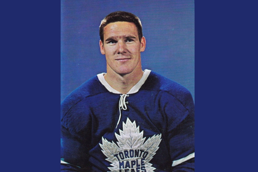 Tim Horton wearing a vintage Toronto Maple Leafs jersey poses in front of a solid blue background. The player is shown from the waist up, dressed in a blue sweater with a large white maple leaf logo and lace-up collar.